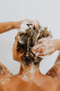 A person with wet, light brown hair, washing their hair with shampoo